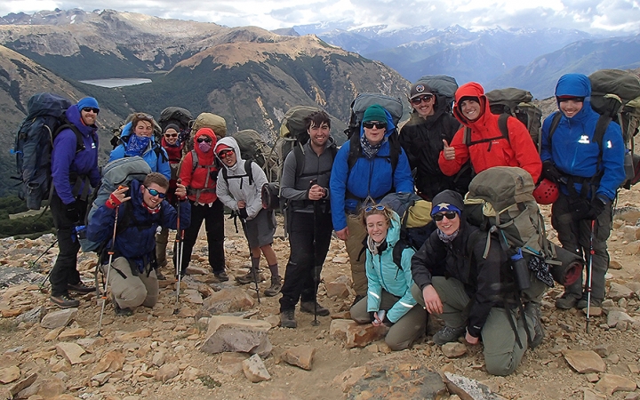 A group of people wearing backpacks pose for a photo while standing on an overlook in front of a vast mountainous landscape.